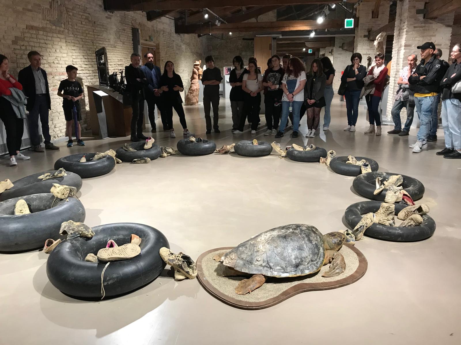L'installazione circolare: a terra a comporre un circolo la tartaruga imbalsamata e 11 pneumatici salvagenti neri su cui poggiano vecchie scarpe infangate.