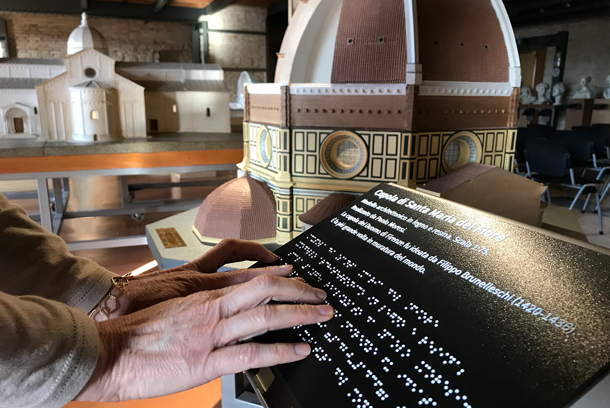 Two hands touch a caption to read Braille. In the background, some wooden architectural models can also be seen.