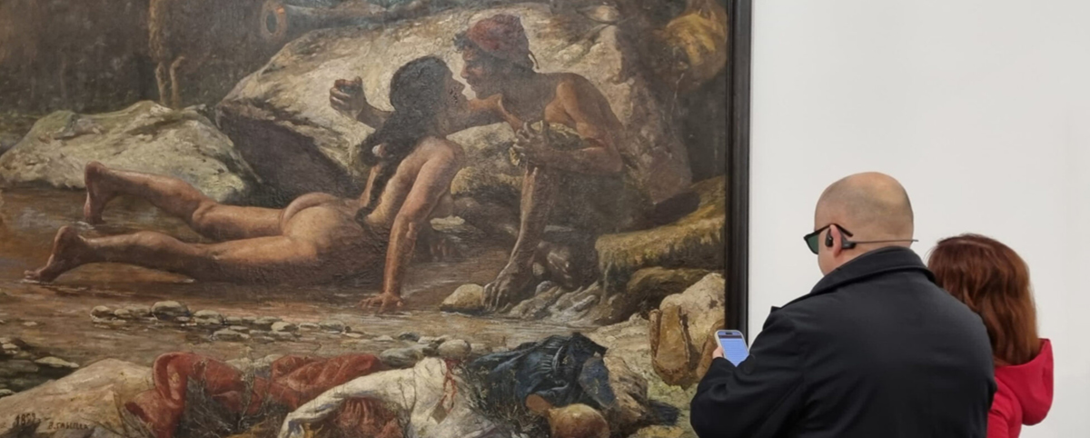 Due visitatori osservano un grande dipinto esposto in museo: una scena naturalistica con due figure umane seminude in un paesaggio roccioso. A destra, un uomo con occhiali scuri e bastone consulta il proprio smartphone, mentre una donna accanto a lui guarda il quadro. L’immagine mette in risalto l’esperienza museale accessibile e l’attenzione all’inclusione.