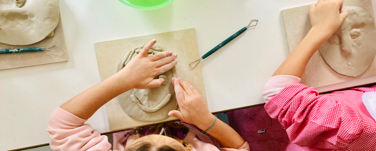 Due bambine modellano volti in argilla su tavolette di legno durante un laboratorio creativo; sulle superfici si vedono strumenti da scultura e piccole ciotole colorate.