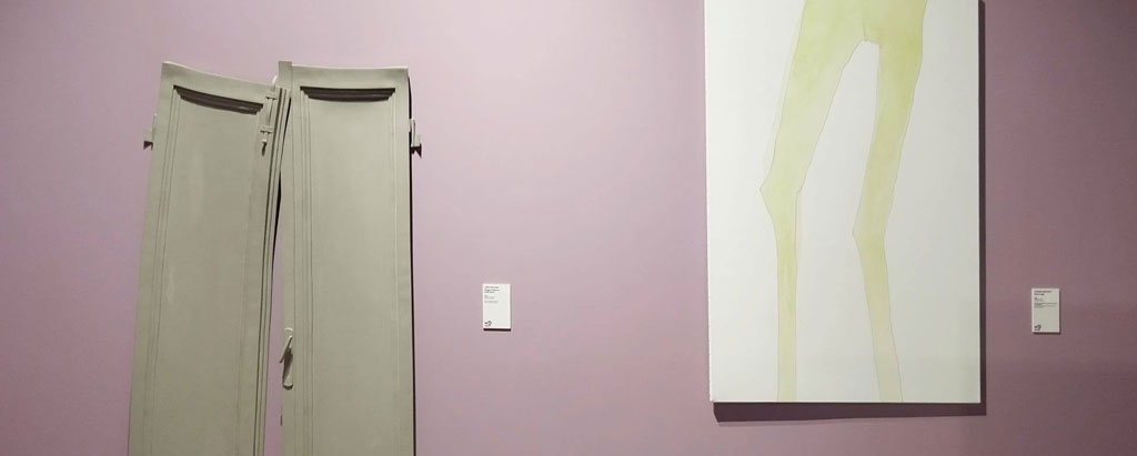 Allestimento museale su parete color malva: a sinistra due ante di legno grigio chiaro, alte e strette, appoggiate verticalmente e deformate; a destra un dipinto su fondo bianco che raffigura solo le gambe di una figura umana, sottili e allungate, dipinte in tonalità giallo-verdastre.