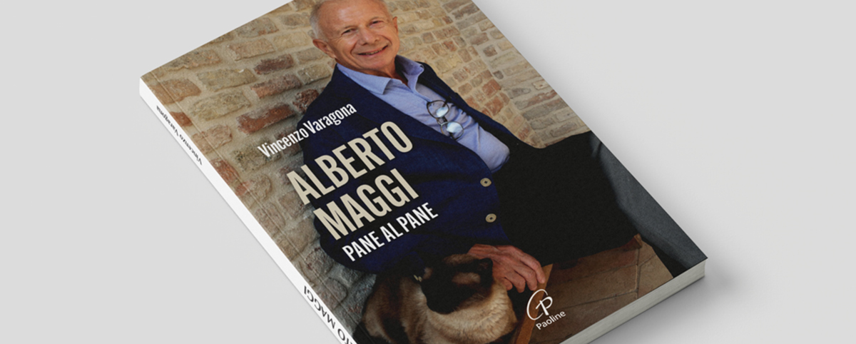 Copertina del libro “Alberto Maggi. Pane al pane” di Vincenzo Varagona: ritratto fotografico di Alberto Maggi sorridente, in piedi davanti a un muro in mattoni chiari, con una giacca scura e una camicia chiara; accanto a lui un gatto seduto. Titolo del libro ben visibile in primo piano. Logo Paoline in basso a destra.