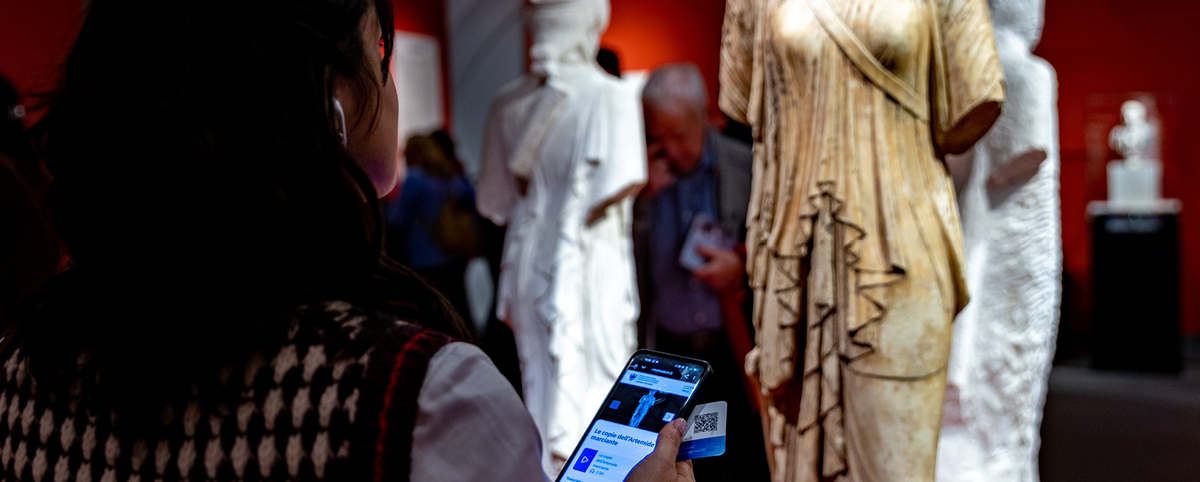 Visitatrice in un percorso museale osserva una scultura classica mentre consulta contenuti accessibili sullo smartphone; sullo sfondo altre statue e persone in mostra.