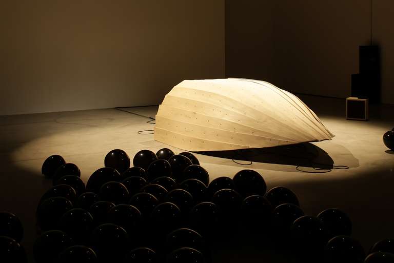 Installazione The Conversation di Francesca Grilli: una grande struttura in legno chiaro, dalla forma che ricorda una conchiglia, è illuminata al centro di una sala. In primo piano, numerosi palloncini neri lucidi sono disposti sul pavimento, pensati per trasmettere vibrazioni sonore attraverso il tatto.