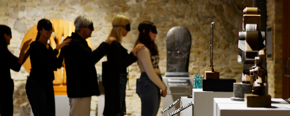 Un gruppo di cinque persone, disposte in fila una dietro l’altra e bendate, esplora lo spazio espositivo del Museo Omero seguendo il contatto delle mani sulle spalle della persona davanti. Accanto a loro sono esposte sculture con didascalie in braille e nero