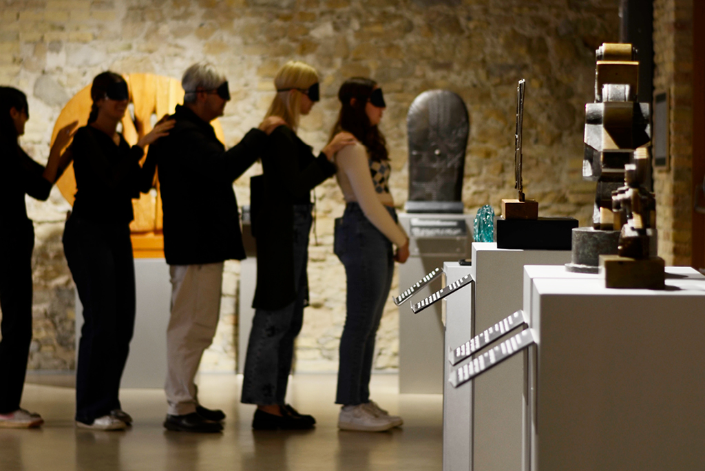 Un gruppo di cinque persone, disposte in fila una dietro l’altra e bendate, esplora lo spazio espositivo del Museo Omero seguendo il contatto delle mani sulle spalle della persona davanti. Accanto a loro, su piedistalli, sono esposte sculture con didascalie in braille e nero