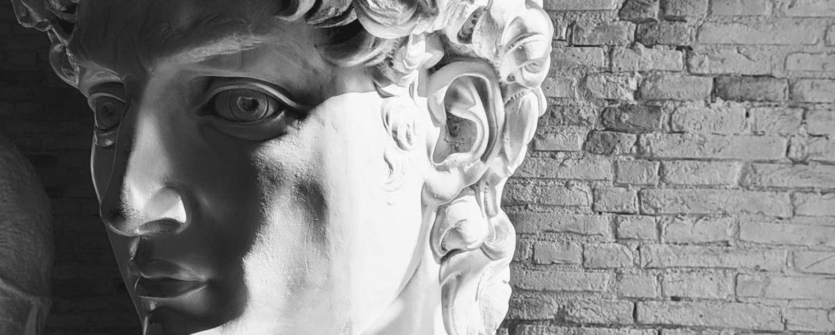 Primo piano del volto del David di Michelangelo, illuminato lateralmente: lo sguardo intenso e i ricci scolpiti emergono dal marmo, mentre sullo sfondo si intravede un muro di mattoni.