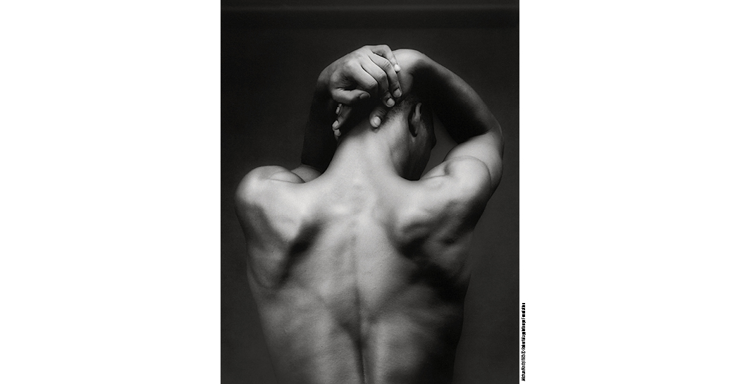 Fotografia in bianco e nero di Robert Mapplethorpe che ritrae la schiena nuda di un uomo vista di spalle. Il corpo emerge dal fondo scuro con forte contrasto chiaroscurale: le scapole, la colonna vertebrale e la muscolatura sono evidenziate dalla luce. Le braccia sono sollevate sopra la testa e le mani si intrecciano dietro il capo.