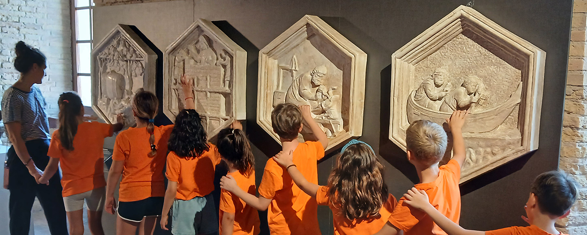 Un gruppo di bambine e bambini, accompagnati da un’educatrice, esplora con le mani alcune riproduzioni in rilievo delle formelle del Campanile di Giotto esposte al Museo Omero.