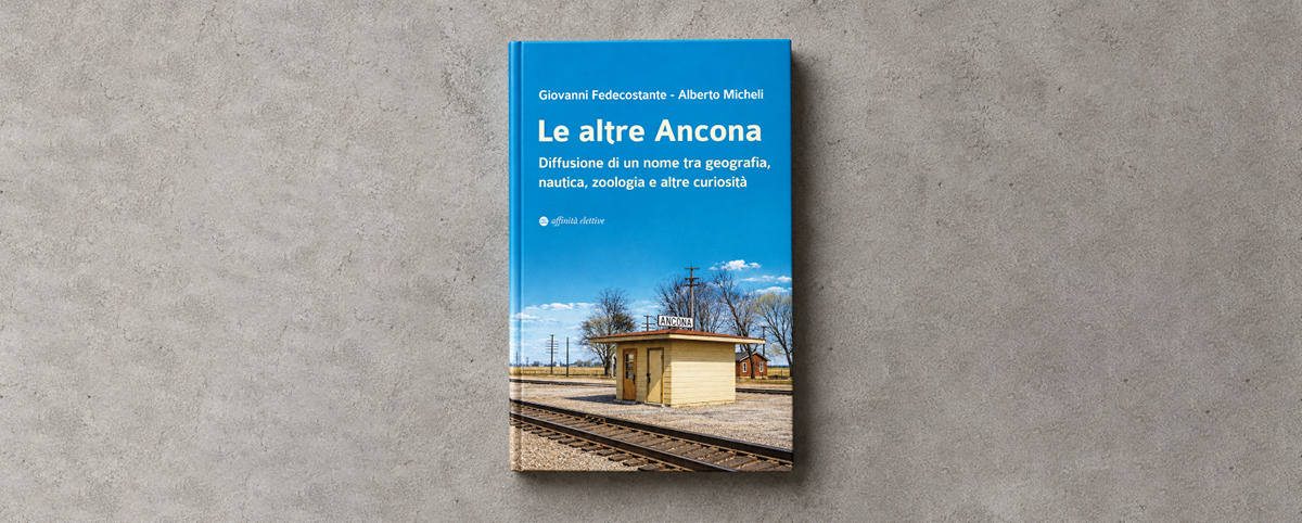 Copertina del libro “Le altre Ancona” di Giovanni Fedecostante e Alberto Micheli, appoggiata su una superficie grigia. La copertina è azzurra, con titolo e nomi degli autori in bianco. Il sottotitolo è “Diffusione di un nome tra geografia, nautica, zoologia e altre curiosità”. Nella parte inferiore compare una fotografia di una piccola stazione ferroviaria in un paesaggio rurale, con un edificio giallo, il cartello “ANCONA” e i binari in primo piano. In basso è presente il logo della casa editrice Affinità Elettive.