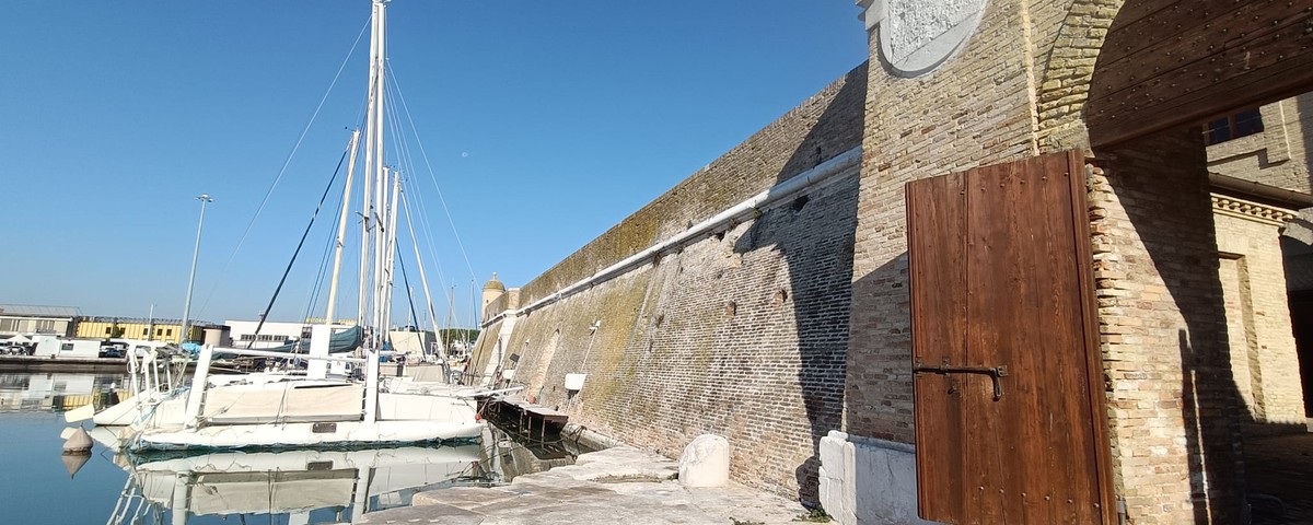 Veduta esterna della Mole Vanvitelliana di Ancona dal lato del porto, con il lungo muro in mattoni e un grande portone in legno; sulla sinistra alcune barche a vela ormeggiate nell’acqua.