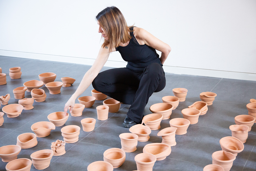 Lucia Cappellazzi, vestita di nero, è accovacciata sul pavimento di uno spazio espositivo mentre dispone piccoli vasi in terracotta di forme e dimensioni diverse. L’artista allunga una mano verso uno dei vasi, distribuiti sul pavimento grigio. L’immagine documenta l’installazione Questo è un orto!, opera finalista della seconda edizione del Premio Omero.